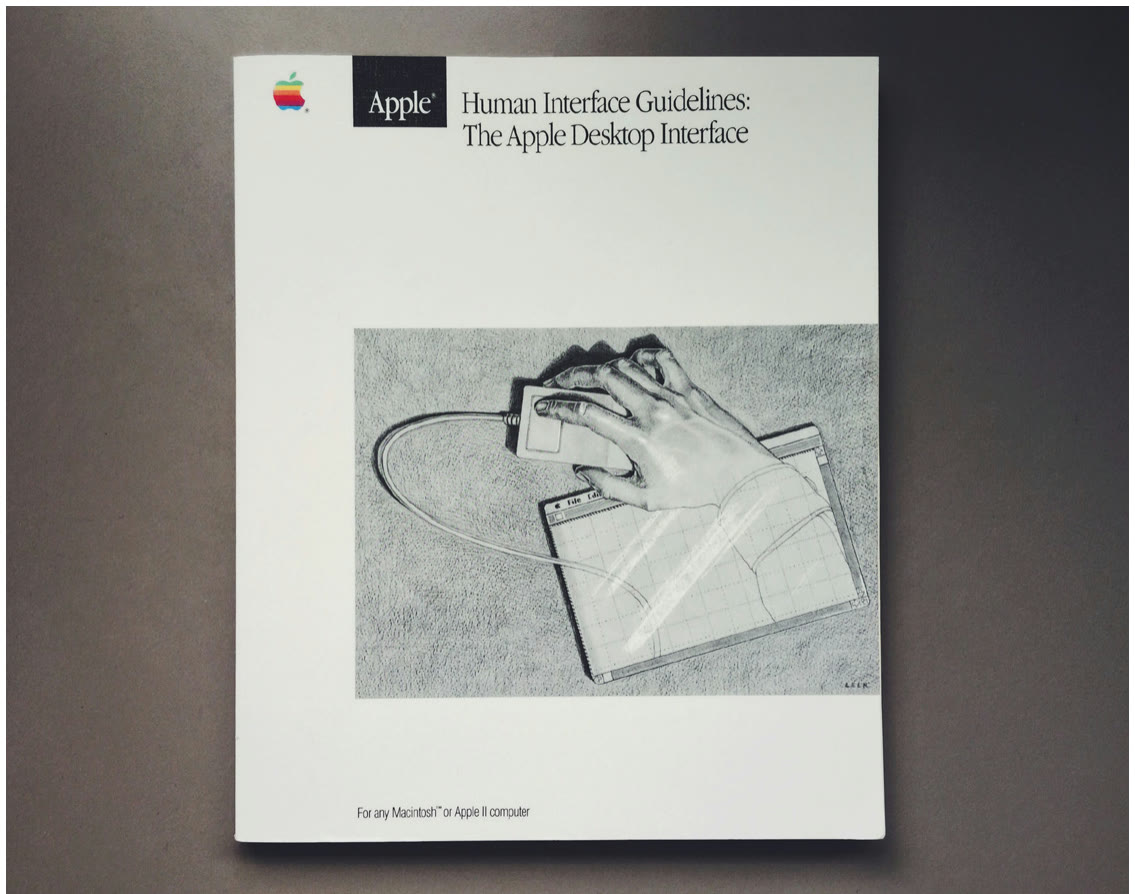 Photo of the printed 1987 Apple Human Interface Guidelines manual, titled The Apple Desktop Interface, held in a person's hand against a dark background.