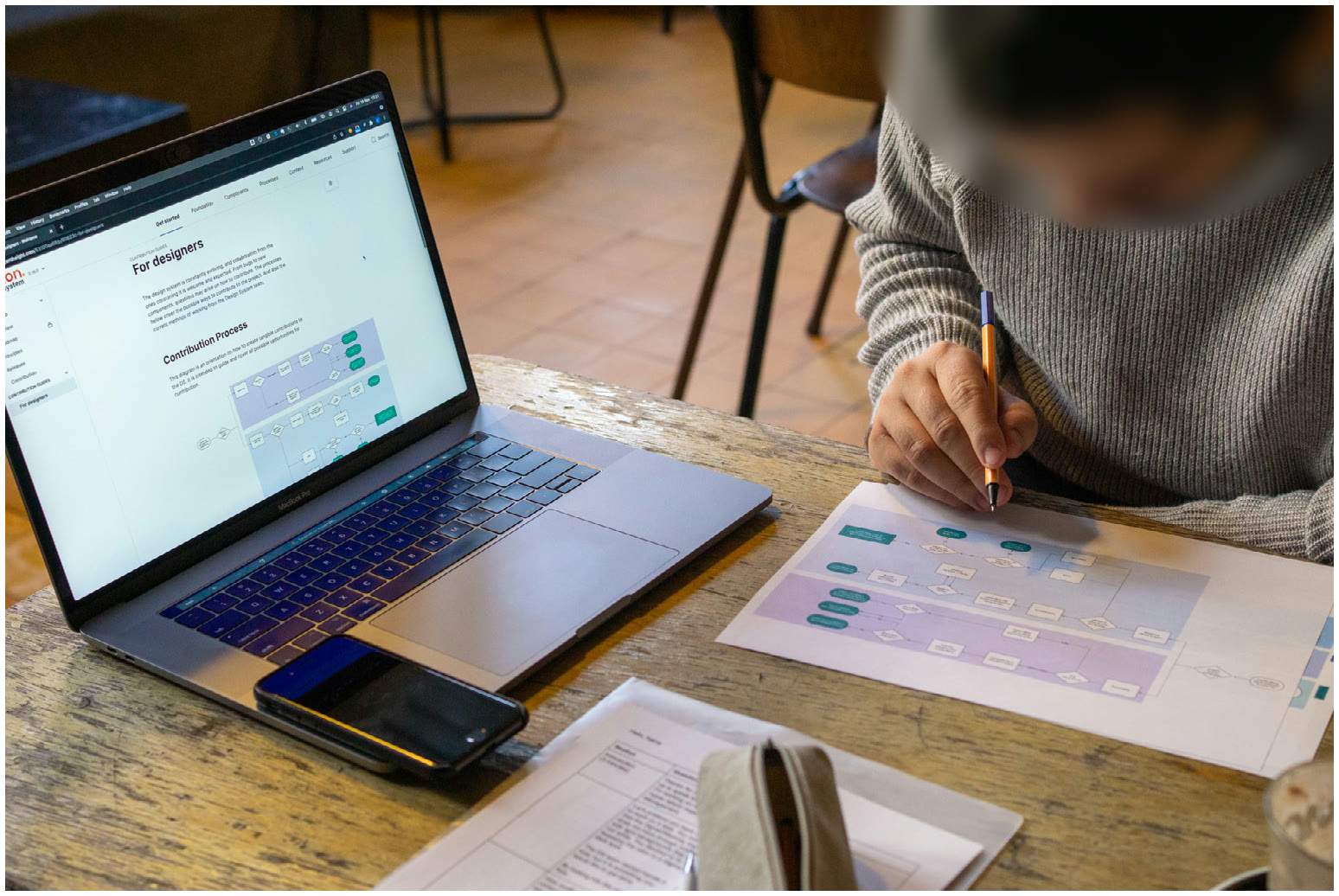 Overhead photo of a person's hand marking up a printed process diagram next to an open laptop showing the HDS documentation, with the participant's face blurred for privacy.